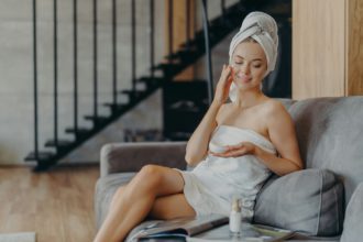 Young female model applies face cream, has perfect body, healthy smooth skin, sits in cozy room on s